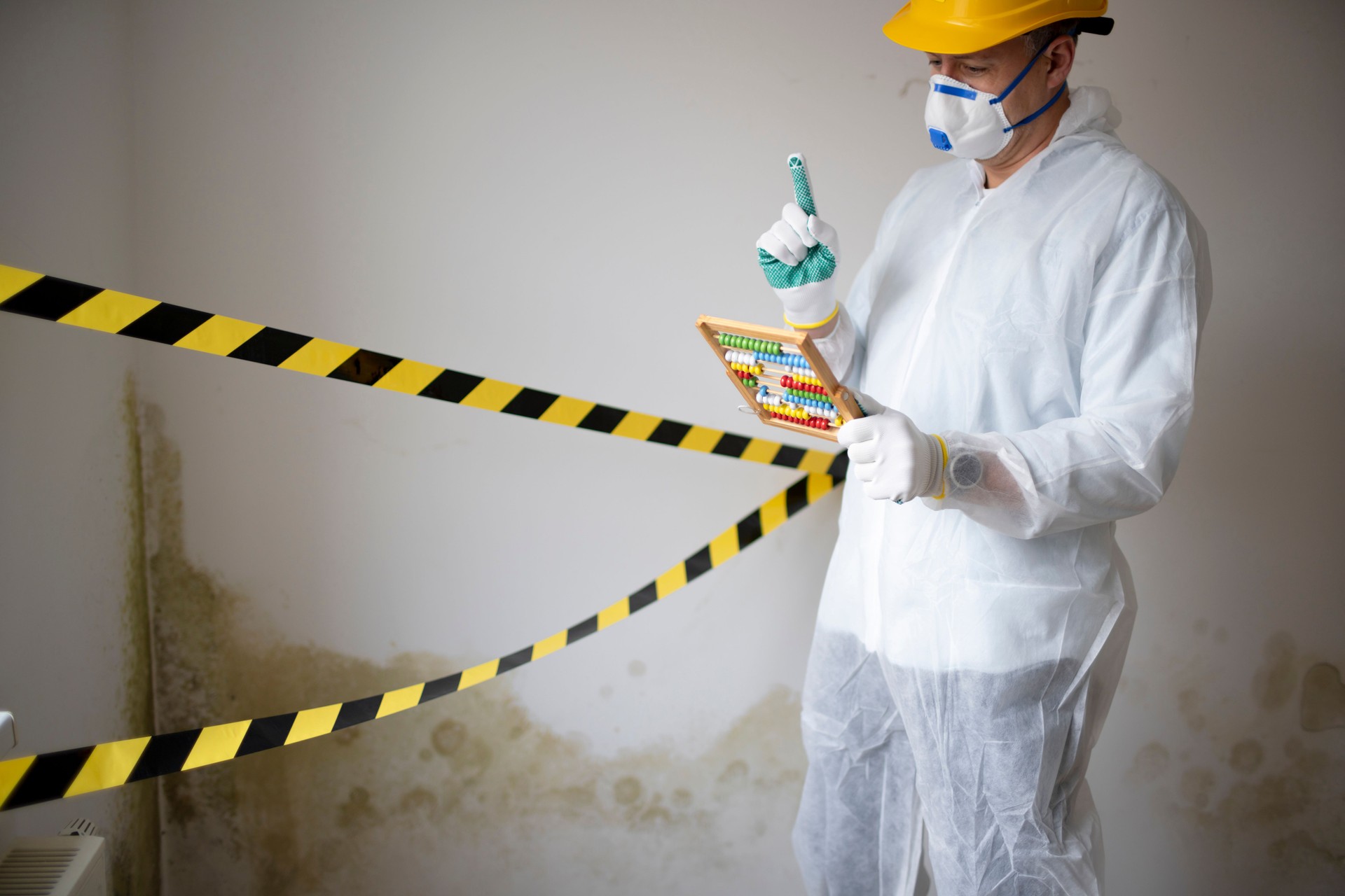 Man with white protective suit with abacus calculator, slide rule in hand and mouth nose mask in front of barrier tape in front of wall with mold
